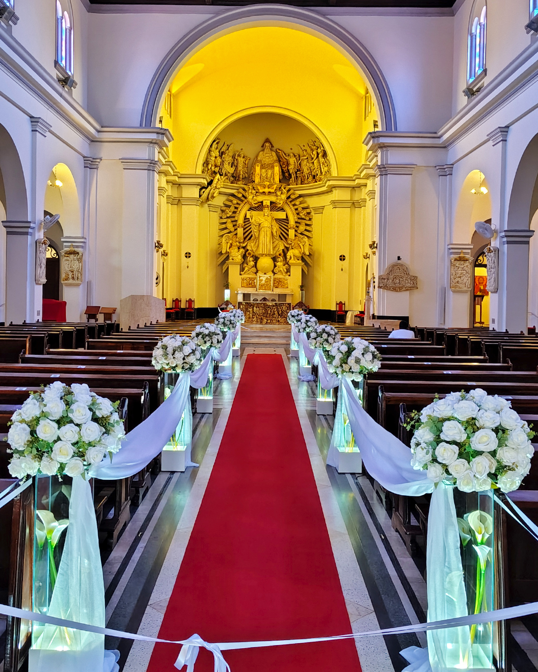 Decoração de cerimônia de casamento na Igreja Cristo Rei zona leste SP com colunas de vidro, tecidos brancos e tapete vermelho no corredor cerimonial